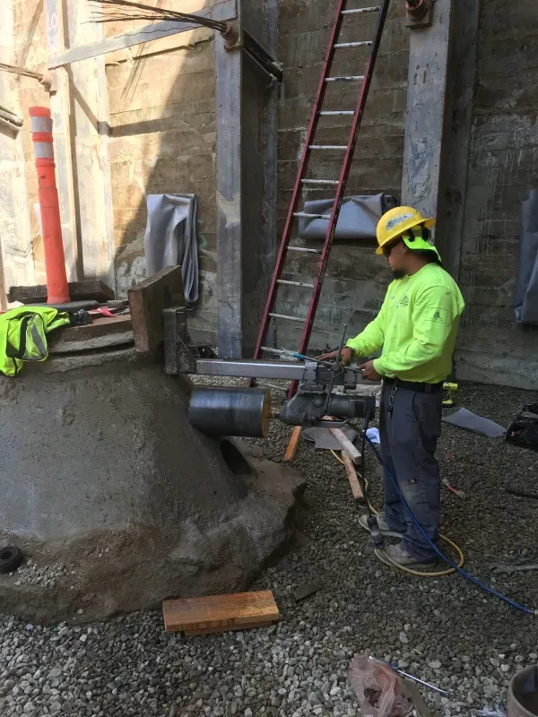 A worker in high-visibility gear uses a diamond core drill on a concrete structure at a construction site.