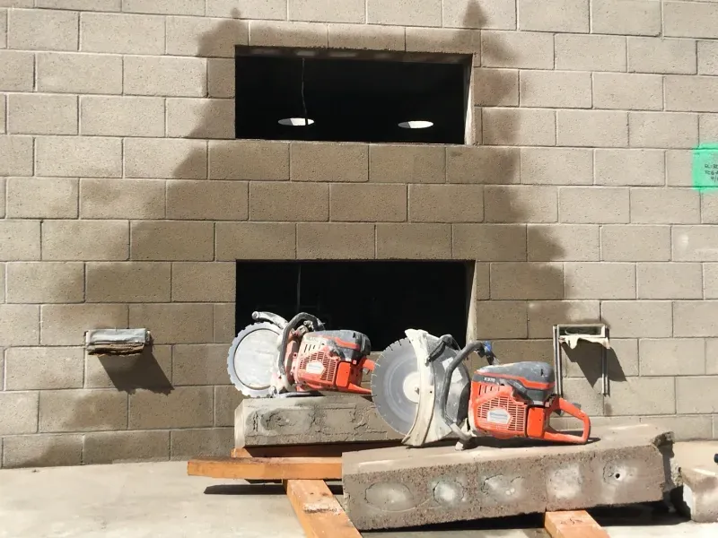 Two orange concrete saws sit on wooden supports in front of a concrete block wall with two rectangular cutouts.
