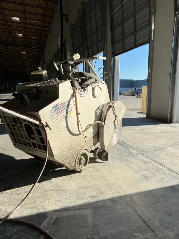 A beige walk-behind concrete saw sits on a dusty warehouse floor near an open bay door.