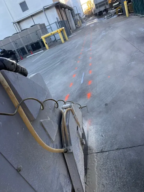 A concrete saw cuts along a path marked with orange spray-paint lines on an industrial parking lot.