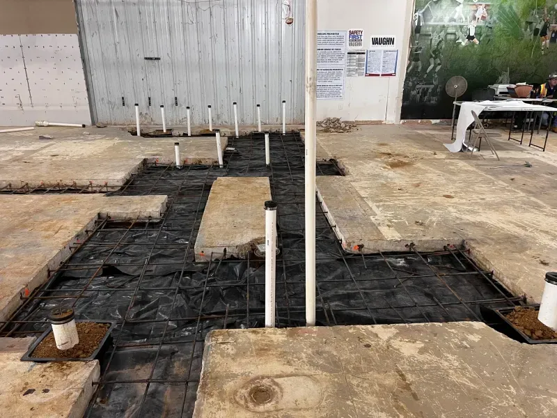 A construction site showing concrete floor sections removed, revealing exposed plumbing pipes and wire mesh reinforcement.