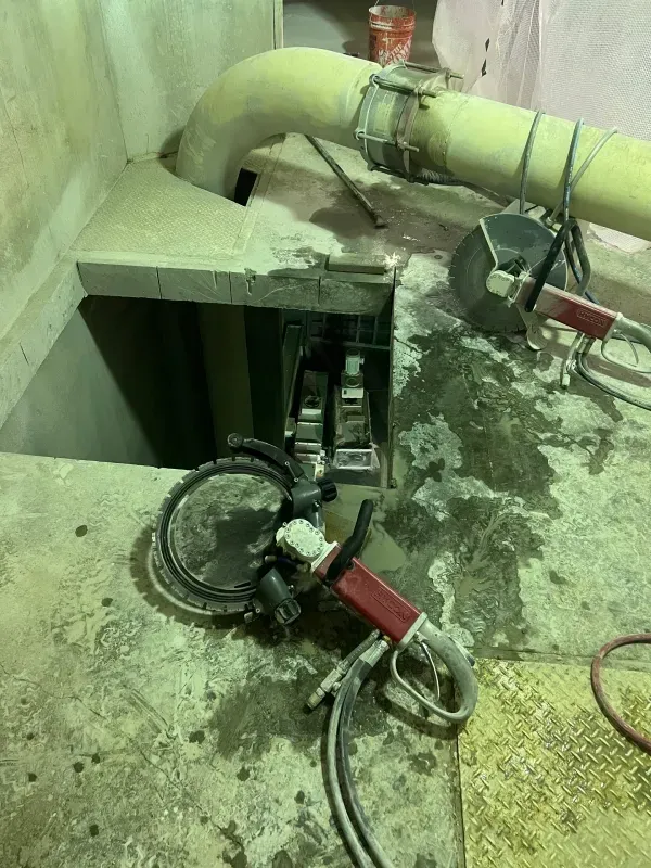 A handheld concrete ring saw lies on a floor near a freshly cut rectangular opening in a concrete slab with a pipe above.