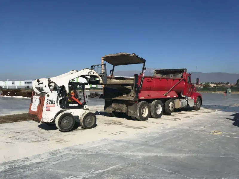 A white skid steer loader parked behind a large red dump truck at an outdoor construction site under a clear blue sky.