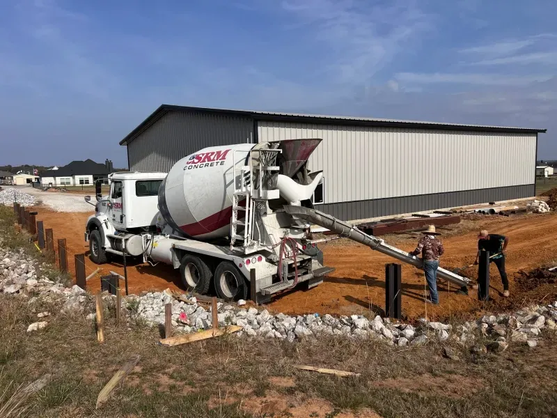 A white concrete mixer truck pours wet cement into a post hole at a construction site near a large metal building.