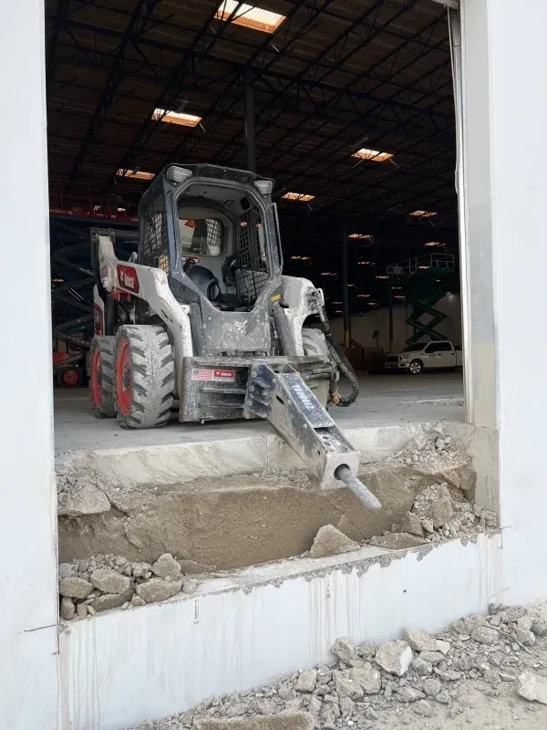 A skid-steer loader with a concrete breaker attachment at the edge of a warehouse loading dock, breaking concrete debris.