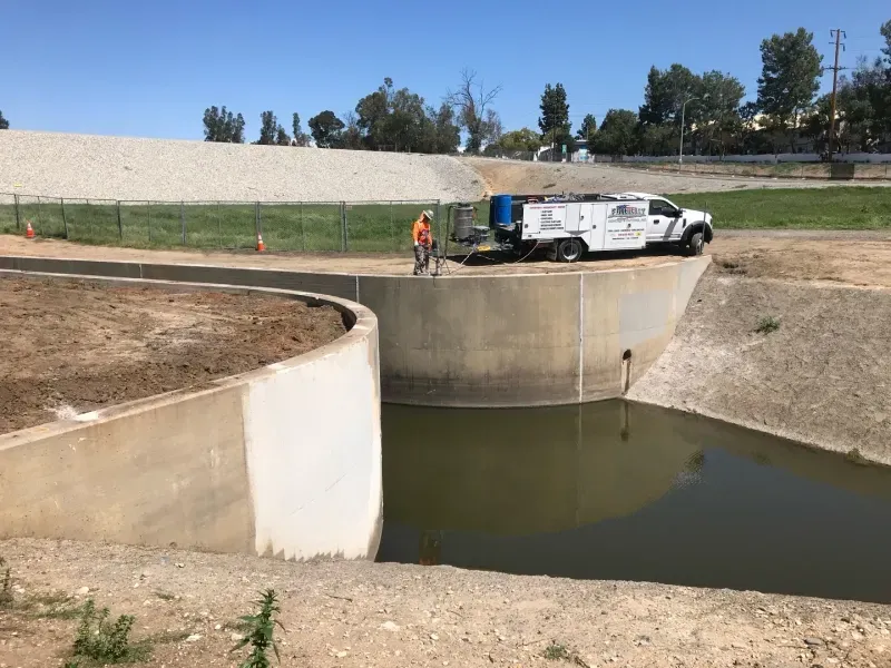 A worker in an orange vest stands near a service truck parked by a concrete storm drain basin filled with murky water.