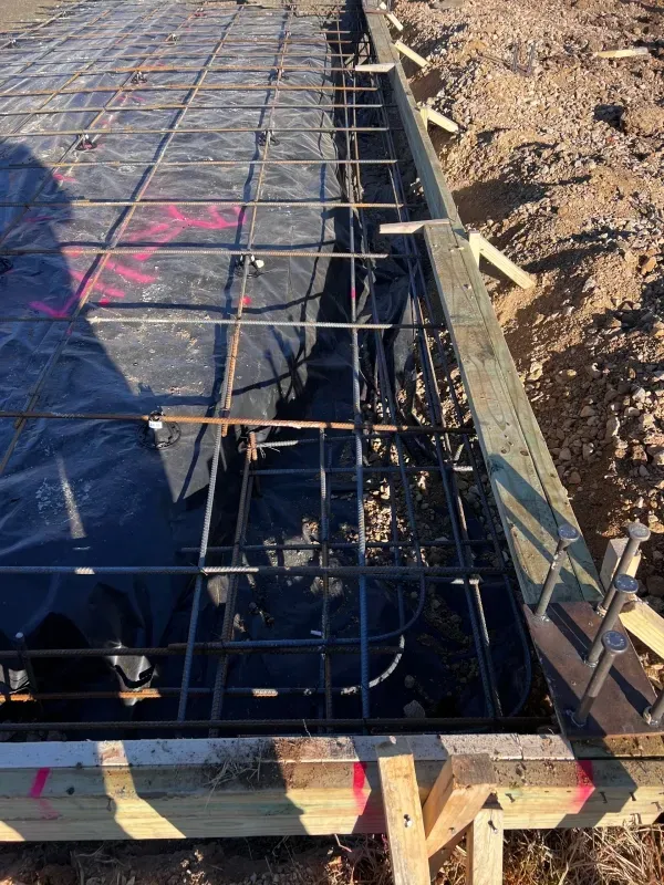 Metal rebar grid laid over a black vapor barrier inside wooden formwork for a concrete foundation pour.
