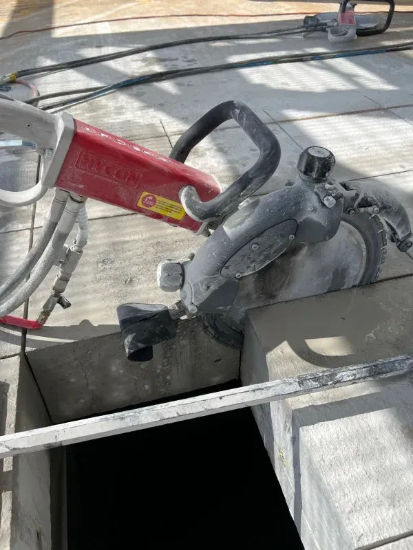 A red industrial power saw cutting a deep rectangular hole in a concrete floor at a construction site.