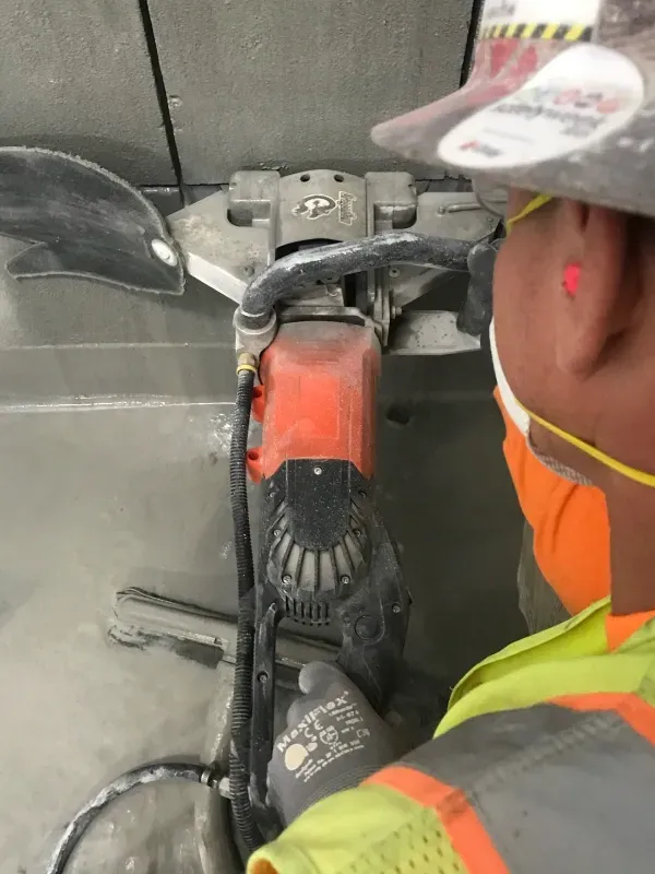 A worker in high-visibility gear uses a handheld concrete saw to cut a line into a gray concrete floor.