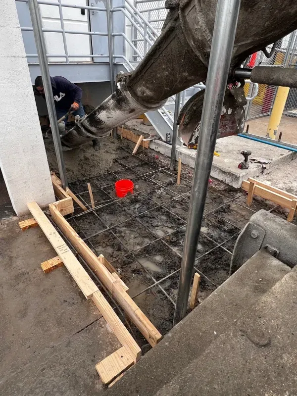 A worker directs wet concrete from a chute into a wooden frame prepared with wire mesh for a small slab.