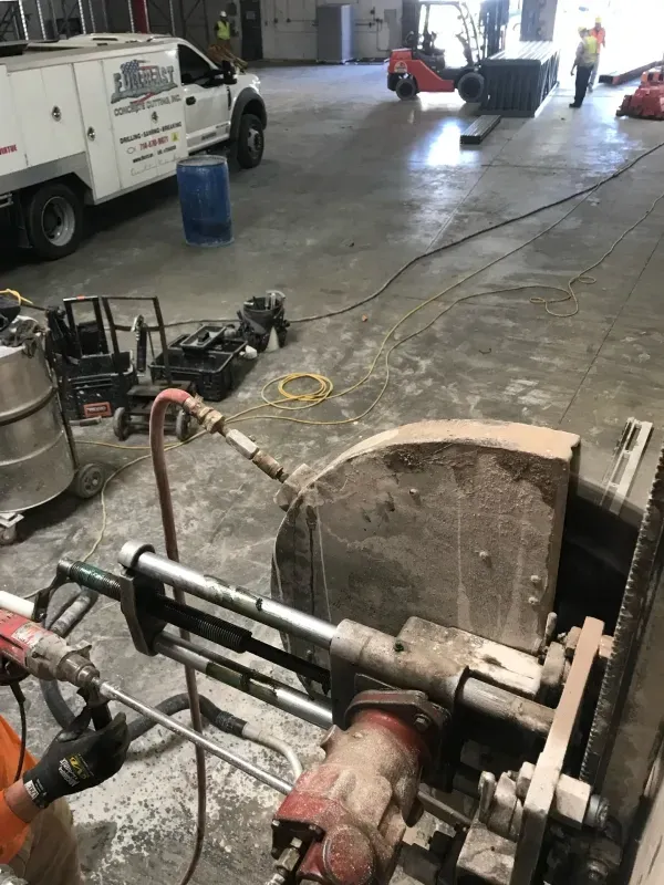 A close-up of a concrete wall saw in a warehouse, with a service truck and a forklift visible in the background.