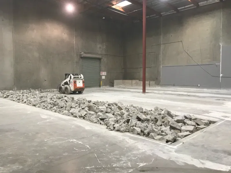 A white skid-steer loader sits in a large warehouse next to a long trench filled with broken concrete rubble.