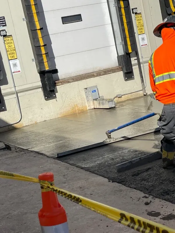 A worker in a high-visibility orange jacket uses a long-handled concrete float to smooth a new surface at a loading dock.