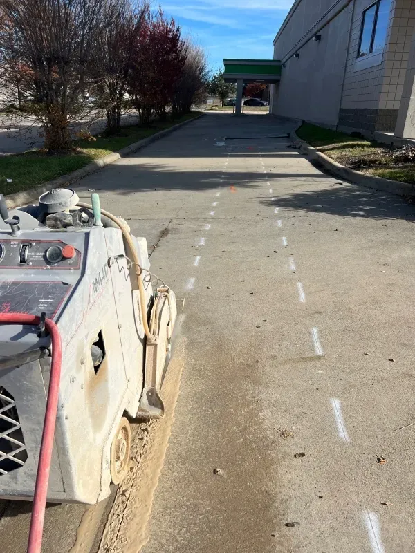 A walk-behind concrete saw cutting a line in an outdoor parking lot marked with white dashed spray paint lines.