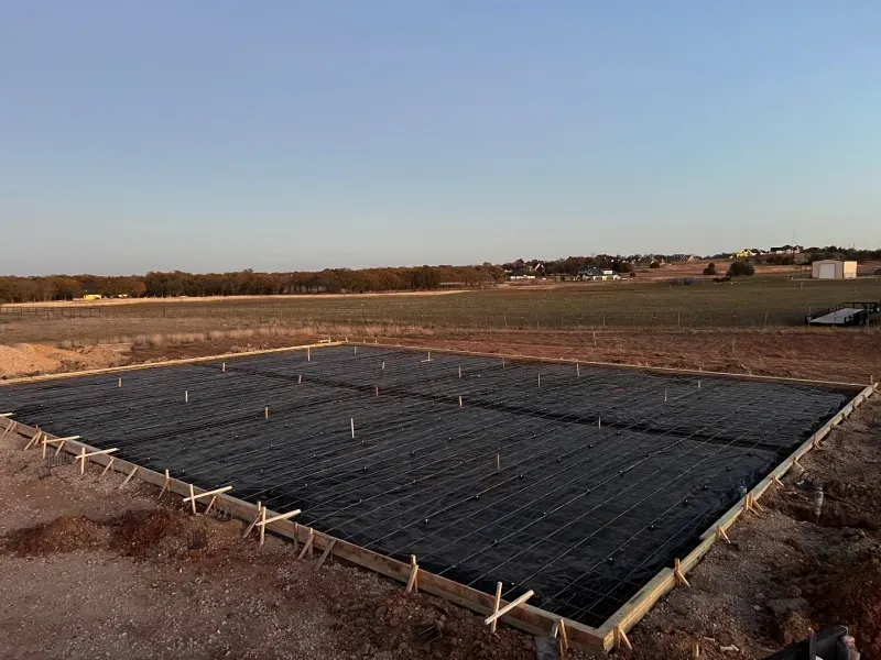 A rectangular concrete slab foundation in a rural field, surrounded by wooden forms and wire mesh reinforcement.