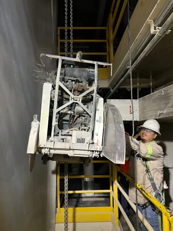 A worker in a hard hat guides a large concrete saw hanging from chains in an industrial setting.