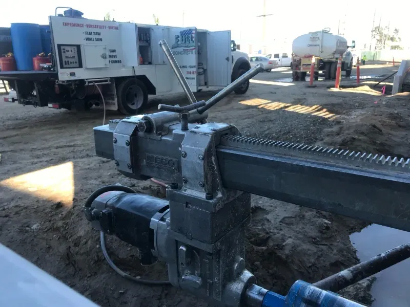 A close-up view of a metal concrete core drill rig at an outdoor construction site, with work trucks in the background.