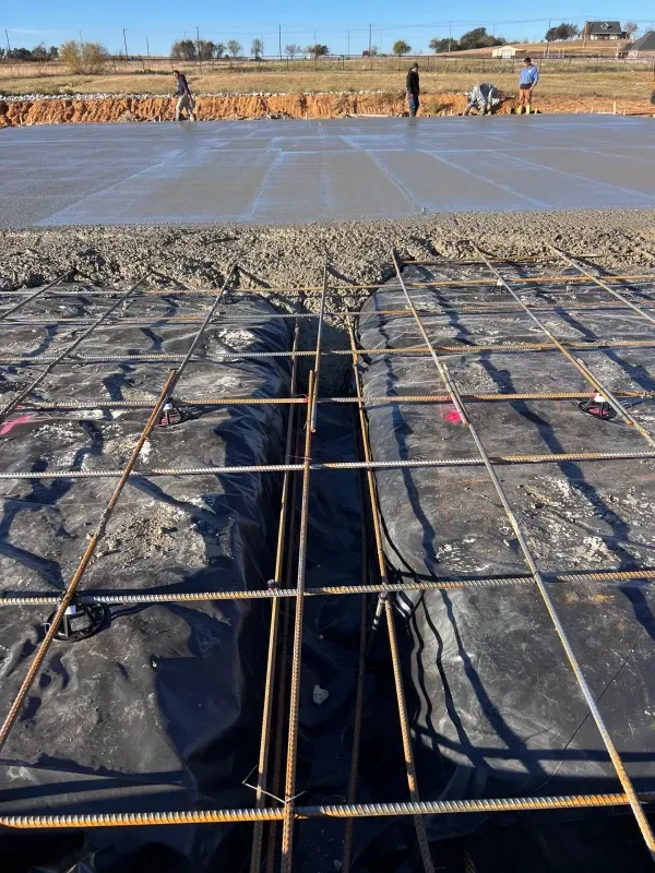 Construction site with rebar grids laid over a vapor barrier in the foreground and wet concrete being poured in the back.