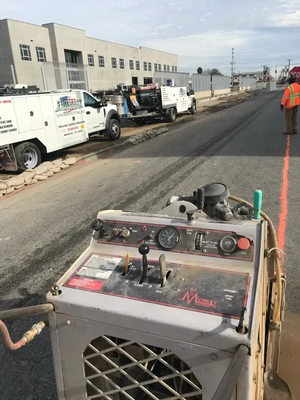 A construction site showing a concrete-cutting machine in the foreground and a work truck and a worker on a road.
