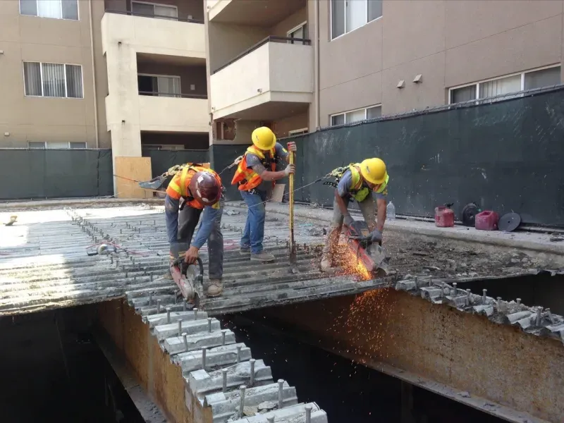 Three construction workers in yellow hard hats and high-visibility vests cut through concrete and metal on a building site.