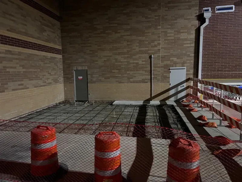 Construction site area with rebar mesh on the ground, orange traffic barrels, and safety fencing against a brick wall.