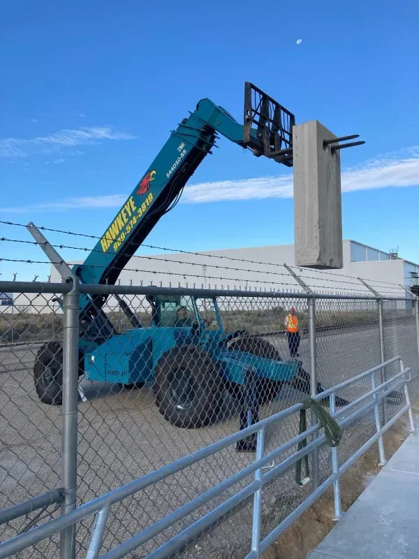 A turquoise telehandler lifts a large concrete barrier over a chain-link fence under a clear blue sky.