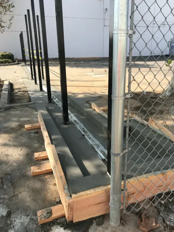 Freshly poured concrete foundation with embedded black metal posts inside wooden formwork near a chain-link fence.