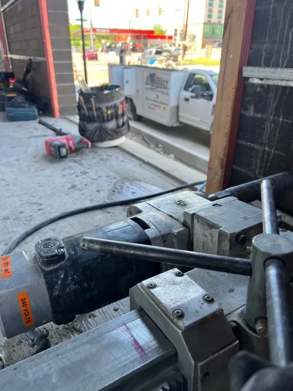 A closeup of a metal industrial core drill machine in a workshop, with a work truck visible through the background window.