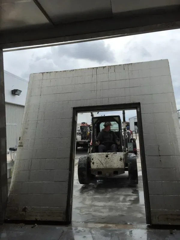 A person in a skid steer loader drives through the doorway of a large, free-standing concrete block wall.