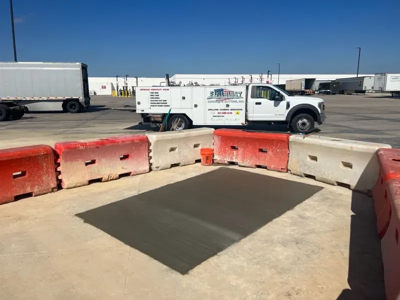 A newly poured concrete patch in a parking lot, surrounded by red and white temporary traffic barriers near a service truck.