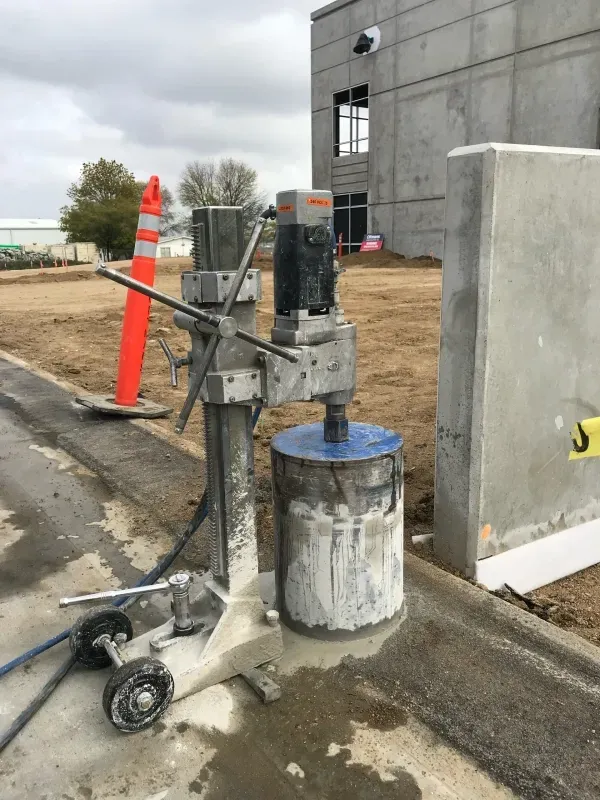 A concrete core drilling machine stands on a construction site next to a concrete wall and an orange traffic cone.