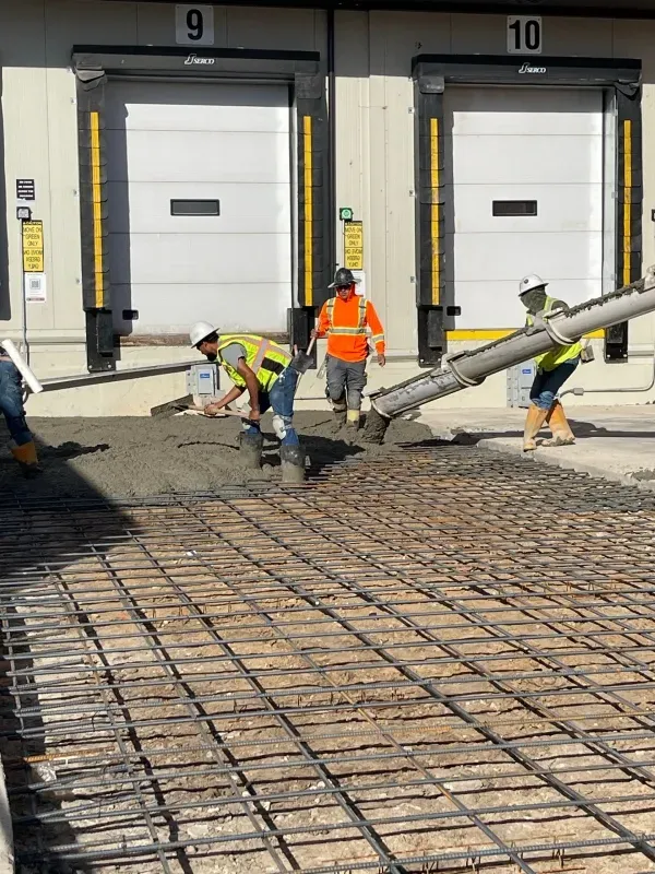 Construction workers pour concrete from a chute over rebar in front of warehouse loading docks marked 9 and 10.