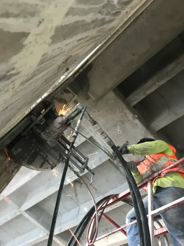A construction worker wearing a high-visibility vest cuts into a concrete ceiling beam, creating sparks.