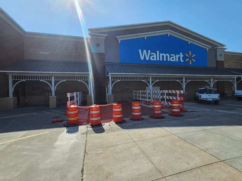 The front entrance of a Walmart store with orange construction barrels and fencing blocking the sidewalk under the awning.