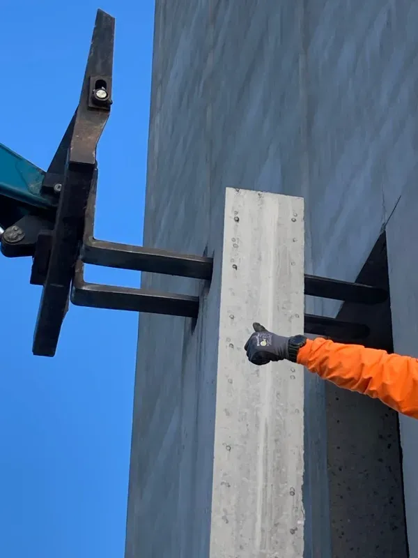 A worker in an orange jacket points to a metal structural support bracket attached to a gray concrete pillar outdoors.