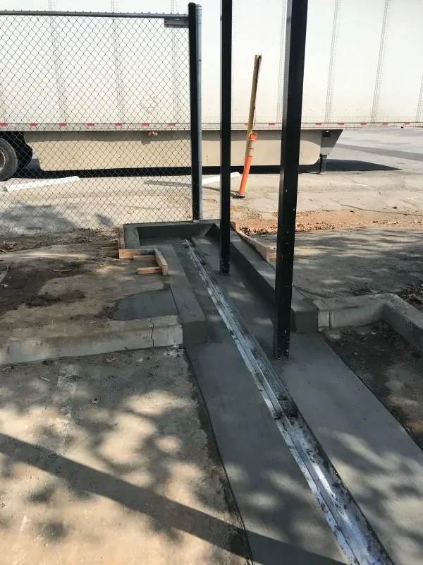 New concrete walkway with an embedded metal track and vertical metal posts installed near a chain-link fence.