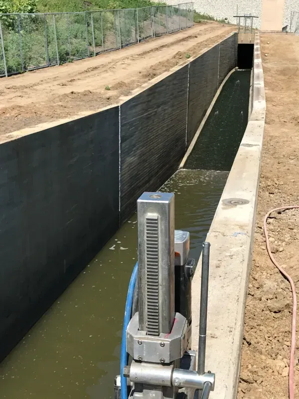 A stainless steel sluice gate mechanism stands in the foreground of a long, concrete water channel in an outdoor area.