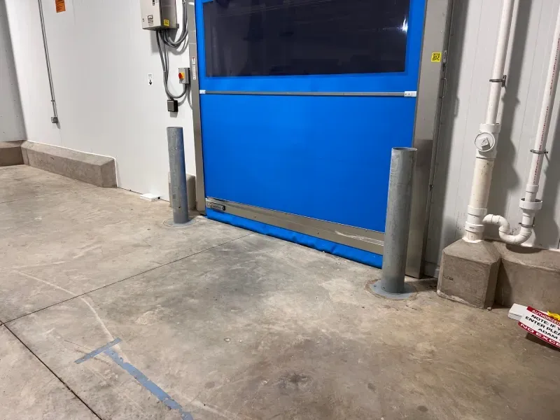 A bright blue industrial high-speed door sits in a gray concrete room, flanked by two protective steel bollards.