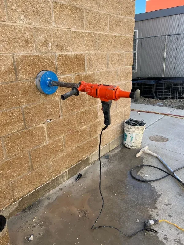 An orange handheld core drill is mounted into a hole being cut into a tan masonry block wall, with a debris bucket nearby.