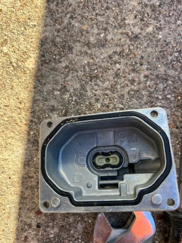 A metallic, rectangular housing component with a black rubber gasket and a central electrical connector, on a concrete surface.