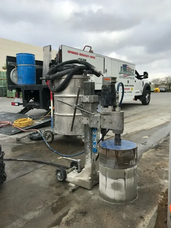 A diamond core drilling rig setup on a concrete surface, with a work truck parked in the background.