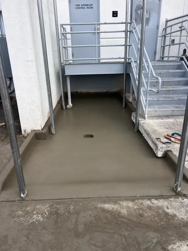 Freshly poured concrete slab beneath metal stairs and a doorway labeled 
