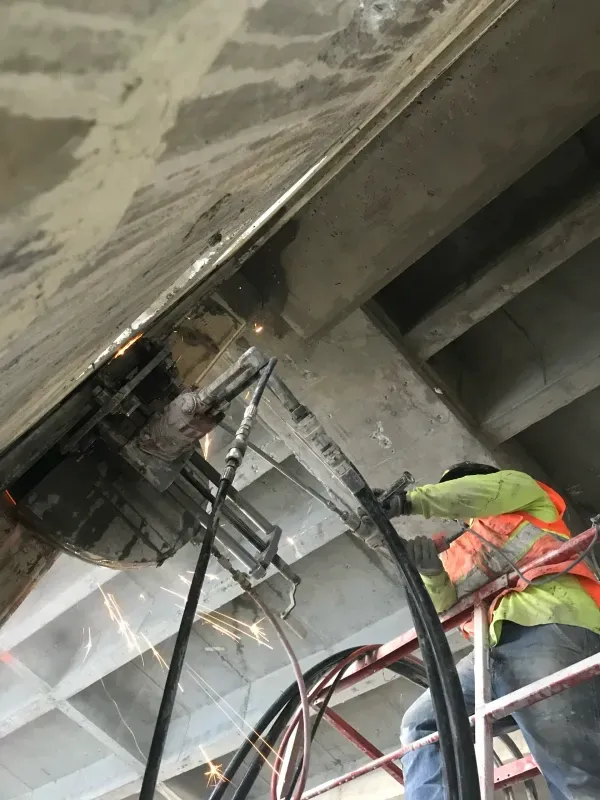 A construction worker on a ladder uses a wall-mounted saw to cut a concrete structure, creating sparks.