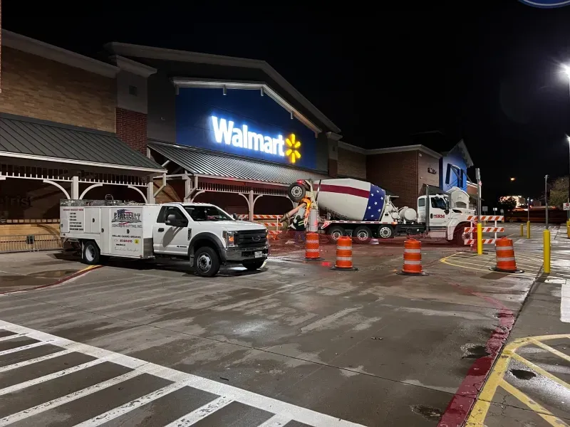 A concrete mixer truck and a service truck parked at a Walmart storefront at night, surrounded by orange traffic cones.