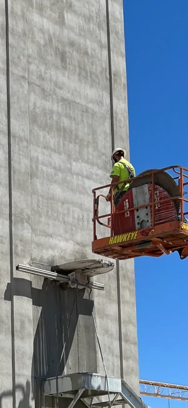 A worker in a high-visibility yellow shirt stands in a red aerial lift, cutting a rectangular hole into a gray wall.