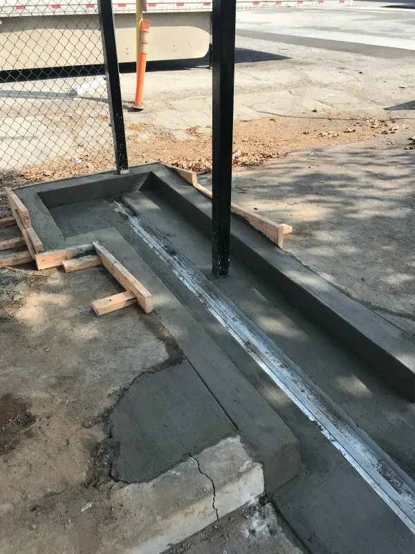 Freshly poured concrete curb and a metal drainage channel installed next to a chain-link fence.