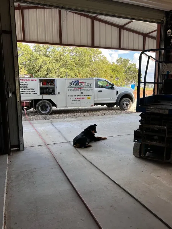 A black dog sits on a concrete shop floor looking toward a white service truck parked outside under a bright sky.