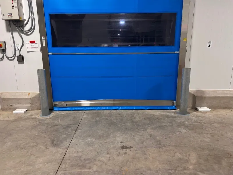 A blue industrial roll-up door with a horizontal window, framed by silver metal posts in a warehouse setting.
