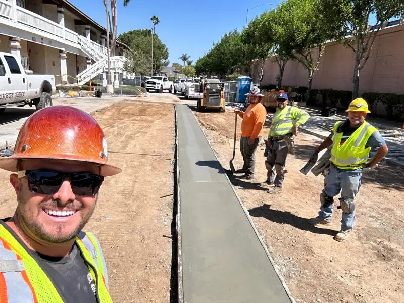 Construction crew in high-vis vests and hard hats stand on a sunny job site next to a freshly poured concrete sidewalk.