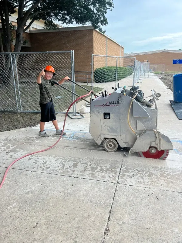 A child in a hard hat gives a thumbs-up while pushing a concrete saw on a sidewalk near a chain-link fence.